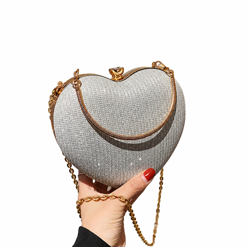 Silver rhinestone heart clutch with gold handle and chain, held in the original hand against a white background.